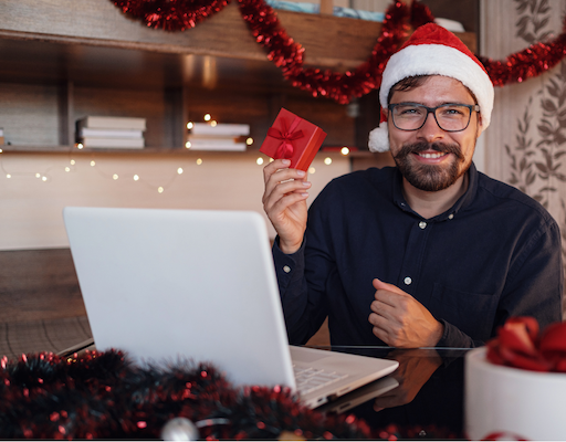 Remote worker in a Santa hat holding a gift at his laptop, illustrating Christmas team activities