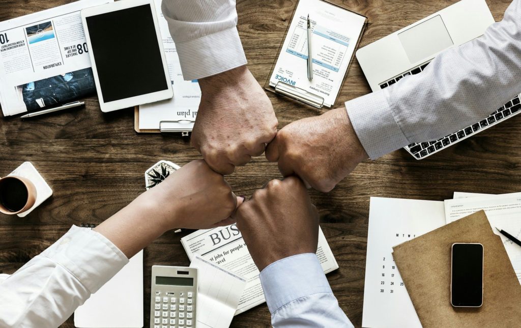 Team members bumping fists over a desk of devices and paperwork, illustrating blended human and tech teams