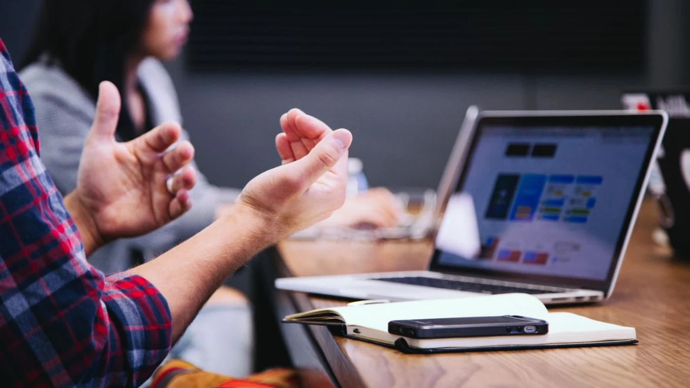 A manager and employee in a one-to-one across a table, the employee gesturing while a laptop and notebook sit between them