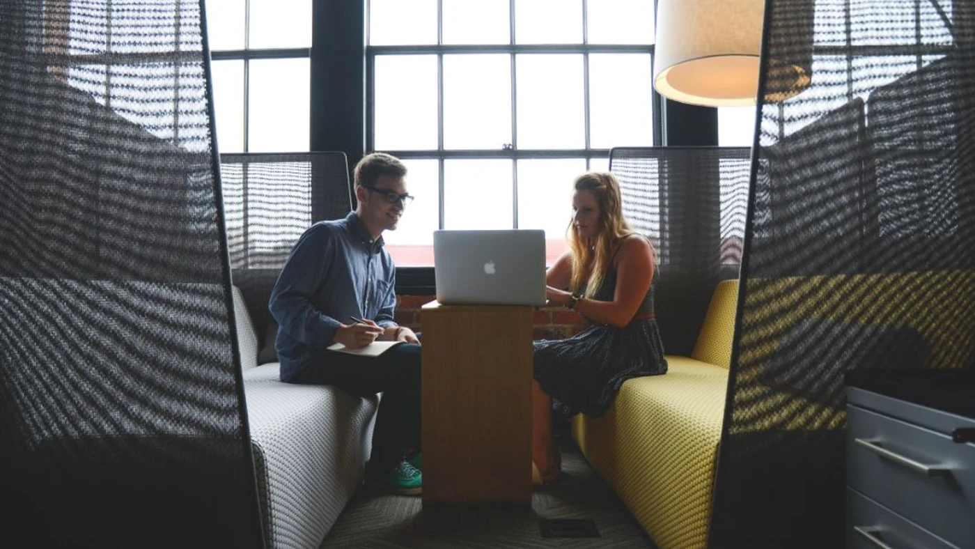 A manager and an employee in a relaxed one-to-one on office sofas by a window, with a laptop open between them